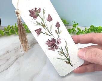 May include: A rectangular white paper bookmark featuring a watercolor illustration of a dark purple flower stem with green leaves. The bookmark has a silver metal eyelet at the top, threaded with a long, light beige decorative silk tassel.