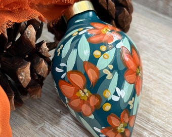 May include: A teardrop-shaped holiday ornament with a teal base color, hand-painted with orange flowers, light green leaves, and small yellow dots. The ornament features a metallic gold-toned cap at the top and a smooth, glossy finish.