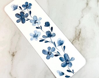 May include: A rectangular bookmark featuring a watercolor painting of a floral vine with various shades of blue flowers and dark blue leaves on a white background. The design displays multiple blossoms arranged along a central stem.