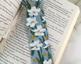 May include: A rectangular bookmark with a dusty blue background featuring hand-painted white flowers with yellow centers, green leaves, and small pink dots. The bookmark includes a white tassel attached to the top and is placed inside an open book.