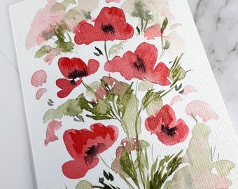 May include: A rectangular greeting card featuring a watercolor painting of vibrant red poppies with dark centers, accented by soft green and pale pink foliage washes on a textured white paper surface, accompanied by a plain white envelope.