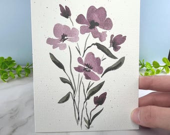 May include: A rectangular greeting card featuring a watercolor illustration of a floral bouquet with mauve-colored petals and dark green stems and leaves. The background of the card is off-white with subtle dark speckles scattered across the surface.