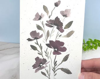 May include: A rectangular greeting card featuring a watercolor illustration of dark purple and muted green flowers on a textured white background. The floral arrangement includes several blossoms, buds, and leaves with scattered paint splatters.