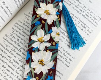 May include: A rectangular wooden bookmark with a dark maroon background featuring hand-painted white flowers with yellow centers, green leaves, and small blue accents. A bright blue silk tassel is attached to the top through a small hole.