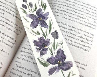 May include: A rectangular paper bookmark featuring a watercolor illustration of three large purple flowers with dark centers and green leaves on a white background. The bookmark is placed on an open book with printed text visible on the pages.