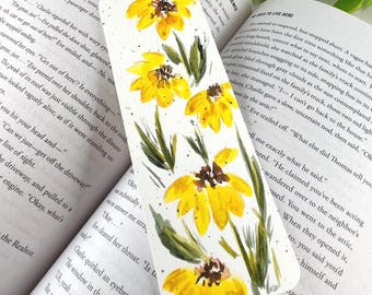 May include: A rectangular bookmark featuring a watercolor painting of bright yellow flowers with dark brown centers and green leaves. The design includes small black paint splatters across the white background, creating a floral pattern.