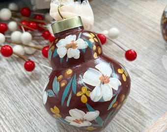 May include: A pear-shaped ornament with a deep brown base color, hand-painted with white flowers, yellow berries, and teal leaves. The ornament features a gold-toned metal cap at the top and is tied with a frayed, cream-colored fabric ribbon.