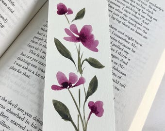 May include: A rectangular paper bookmark featuring a watercolor painting of a vertical floral stem with four purple blossoms and several dark green leaves. The bookmark is placed against the open pages of a printed book with visible text.