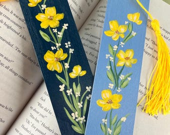 May include: Two rectangular wooden bookmarks, one dark navy blue and one light blue, each hand-painted with yellow flowers, green leaves, and small white blossoms. Both bookmarks feature a hole at the top with a bright yellow tassel attached.