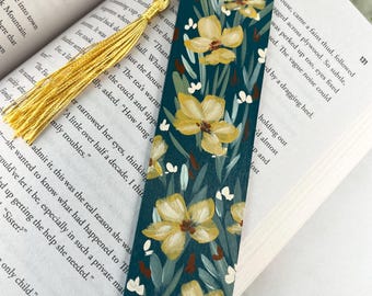 May include: A rectangular bookmark featuring a dark teal background with a painted pattern of yellow flowers, green leaves, and small white accents. The bookmark includes a gold-colored tassel attached to the top with a small metal loop.