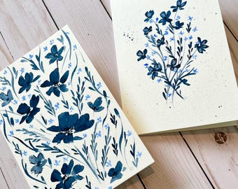 May include: Two rectangular greeting cards featuring hand-painted floral designs in shades of navy and light blue on a cream-colored background. One card displays a dense, scattered pattern of flowers and leaves, while the other shows a central bouquet.