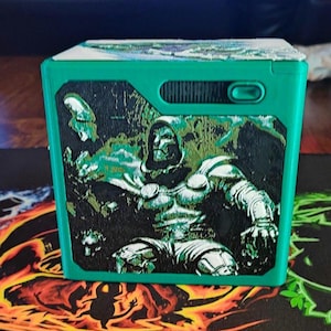 May include: A bright green square box featuring a detailed black and white graphic of a hooded figure in armor on the front panel. The box has a small rectangular opening near the top right corner and a textured surface on the illustrated section.