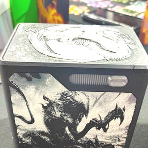 May include: A black rectangular deck box featuring a white embossed dragon emblem on the top lid and a detailed black and white graphic illustration of a creature with sharp claws and a tail on the front panel, alongside a small silver button.