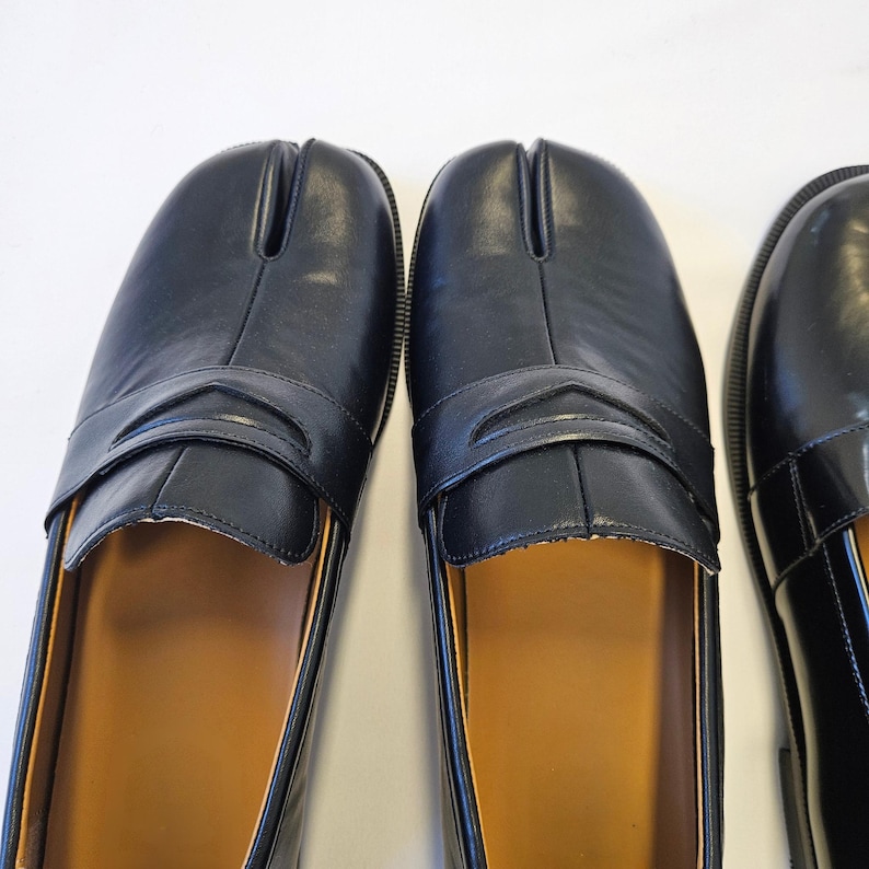 Puede incluir: Un par de mocasines de cuero negro con un dise&ntilde;o distintivo de punta dividida, a menudo llamado estilo tabi. Los zapatos tienen una tira cl&aacute;sica de mocas&iacute;n sobre el empeine, un acabado liso y un forro interior de color canela visible.