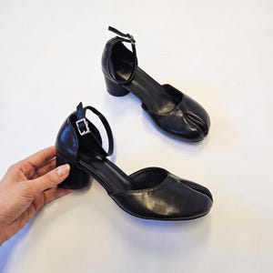 Könnte beinhalten: Ein Paar schwarze Schuhe mit geteilter Zehenpartie, ausgestattet mit einem Blockabsatz und einem verstellbaren Knöchelriemen mit silberfarbener Metallschnalle. Die Schuhe haben eine glatte Oberfläche und ein tief ausgeschnittenes Seitenprofil.