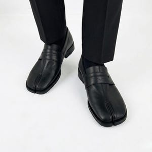 Puede incluir: Un par de mocasines de cuero negro con un dise&ntilde;o distintivo de punta dividida, una correa cl&aacute;sica de mocas&iacute;n sobre el empeine y un tac&oacute;n bajo. Los zapatos se usan con pantalones negros y calcetines negros, mostrando un acabado liso y estructurado.