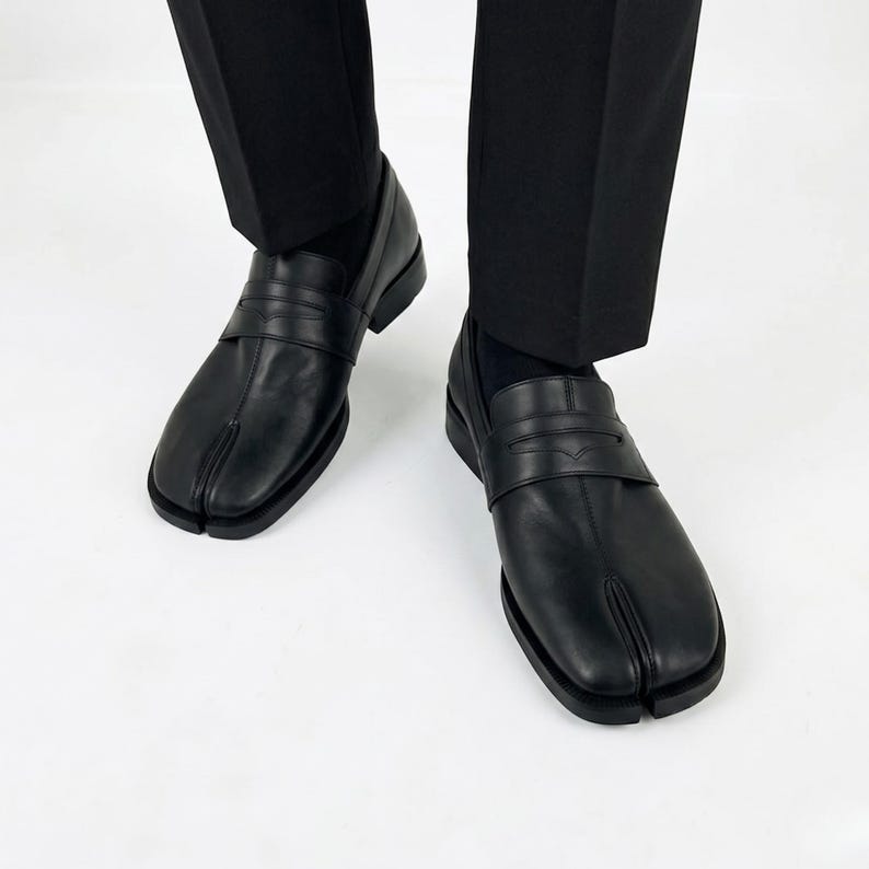 Puede incluir: Un par de mocasines de cuero negro con un dise&ntilde;o distintivo de punta dividida, una correa cl&aacute;sica de mocas&iacute;n sobre el empeine y un tac&oacute;n bajo. Los zapatos se usan con pantalones negros y calcetines negros, mostrando un acabado liso y estructurado.