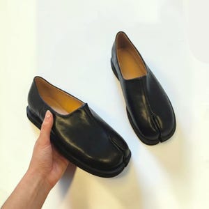 Puede incluir: Un par de zapatos sin cordones de cuero negro con un diseño distintivo de punta dividida en la parte delantera. Los zapatos tienen un acabado mate liso, una silueta de perfil bajo y una suela negra gruesa, con forro interior color canela.