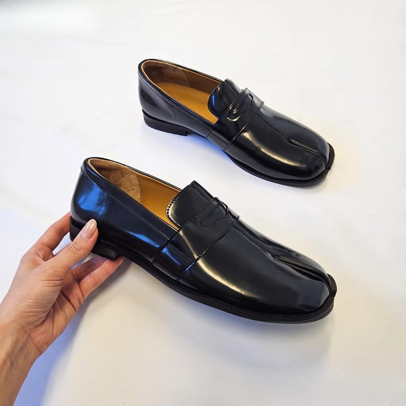 Puede incluir: Un par de mocasines de cuero negro brillante con un dise&ntilde;o distintivo de punta dividida, una tira cl&aacute;sica de mocas&iacute;n sobre el empeine, un tac&oacute;n bajo apilado y un forro interior color canela, presentados como calzado sin cordones.