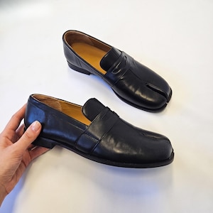Puede incluir: Un par de mocasines de cuero negro con un dise&ntilde;o distintivo de punta dividida en la parte delantera. Los zapatos tienen un acabado liso, una tira cl&aacute;sica de mocas&iacute;n sobre el empeine, un tac&oacute;n bajo y un forro interior de color beige.