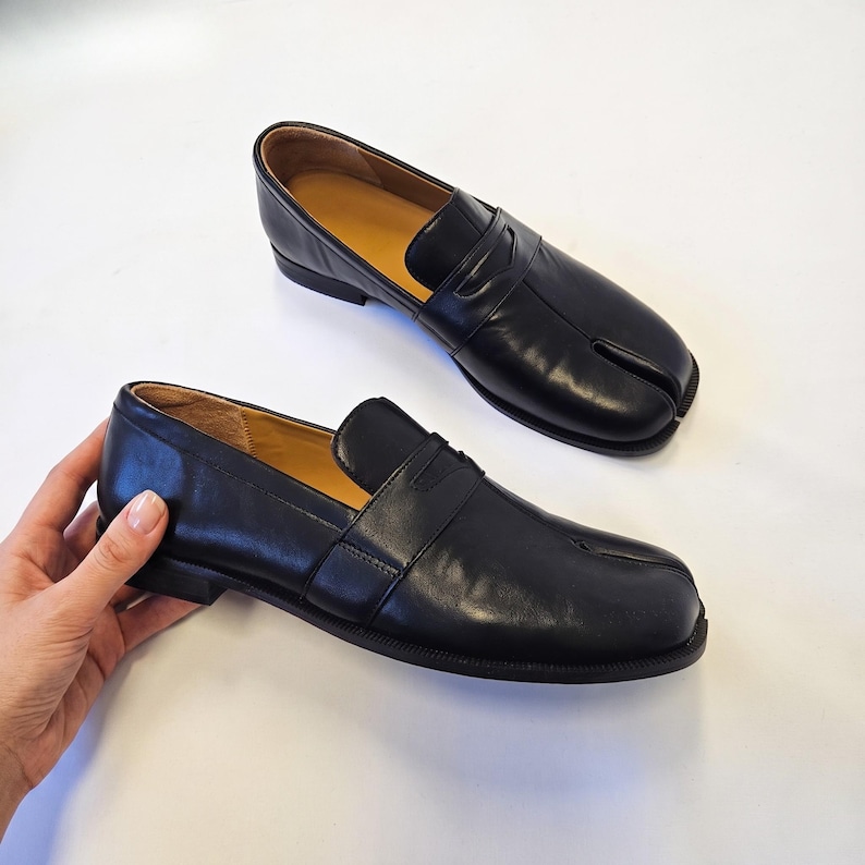 Puede incluir: Un par de mocasines de cuero negro con un dise&ntilde;o distintivo de punta dividida en la parte delantera. Los zapatos tienen un acabado liso, una tira cl&aacute;sica de mocas&iacute;n sobre el empeine, un tac&oacute;n bajo y un forro interior de color beige.