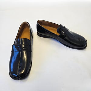 Puede incluir: Un par de mocasines de cuero negro brillante con un dise&ntilde;o distintivo de punta dividida. Estos zapatos sin cordones tienen una tira cl&aacute;sica de mocas&iacute;n sobre el empeine, un tac&oacute;n bajo y un forro interior color canela, con un acabado pulido.