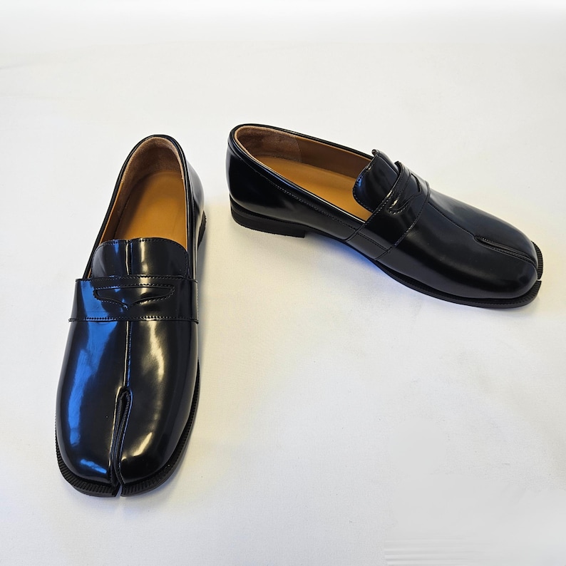 Puede incluir: Un par de mocasines de cuero negro brillante con un dise&ntilde;o distintivo de punta dividida. Estos zapatos sin cordones tienen una tira cl&aacute;sica de mocas&iacute;n sobre el empeine, un tac&oacute;n bajo y un forro interior color canela, con un acabado pulido.