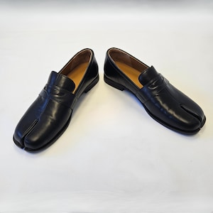 Puede incluir: Un par de mocasines de cuero negro con un dise&ntilde;o distintivo de punta dividida en la parte delantera. Estos zapatos sin cordones tienen una tira cl&aacute;sica de mocas&iacute;n sobre el empeine, un acabado liso y un tac&oacute;n bajo, con un aspecto estructurado.