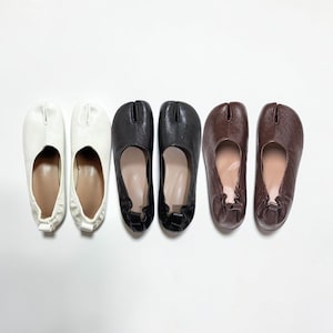 Könnte beinhalten: Drei Paar Schuhe mit geteilter Zehenpartie in Weiß, Schwarz und Braun. Jedes Paar verfügt über ein markantes Schlitzdesign an der Zehenkappe, einen tiefen Ausschnitt und einen gerafften elastischen Fersenbereich. Die Oberfläche ist glatt.