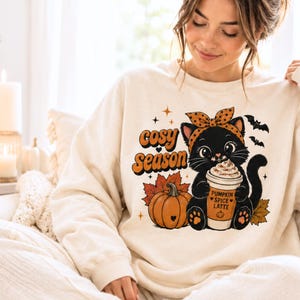 May include: A cream-coloured crewneck sweatshirt featuring a graphic print of a black cat wearing an orange polka-dot headband. The cat holds a cup labelled Pumpkin Spice Latte next to a pumpkin and autumn leaves, with the text Cosy Season above.