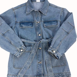 Op de afbeelding: Een medium-wash blauwe spijkerjas met een klassieke kraag, lange mouwen met knoopmanchetten en een bijpassende stoffen ceintuur in de taille. De jas heeft twee borstzakken met kleppen en twee opgestikte zakken, allemaal voorzien van zilverkleurige knopen.