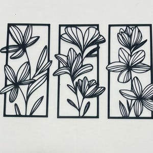 Op de afbeelding: Een set van drie rechthoekige zwarte wandkunstpanelen, elk met een opvallend lineair uitgesneden ontwerp van bloemen en bladeren. De frames zijn dun en solide en omsluiten de botanische illustraties die iets buiten de randen uitsteken.