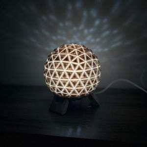 Op de afbeelding: Een bolvormige lamp met een complexe geometrische roosterstructuur bestaande uit onderling verbonden driehoeken. De interne lichtbron verlicht het witte frame en werpt een zachte, patroonachtige gloed op de omgeving. De lamp rust op een donkere voet.