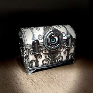 Puede incluir: Un cofre blanco con detalles de filigrana en plata metálica y un elemento circular central que contiene un ojo azul realista. La caja tiene una tapa curva y un adorno decorativo de aspecto metálico en la parte frontal.