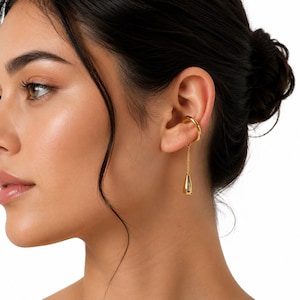 May include: A gold-toned ear cuff featuring a smooth, curved band that wraps around the cartilage. A delicate gold chain hangs from the cuff, ending in a polished, teardrop-shaped pendant that rests against the earlobe area.