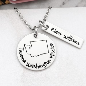 May include: A silver necklace featuring two pendants. One is a circular pendant with the outline of Washington state and the words "Tacoma Washington Mission". The other is a rectangular pendant with the words "Elder Williams".