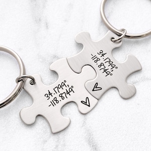 May include: Two silver puzzle piece keychains connected, each with a keyring. One piece has the coordinates 34.1799°, -118.8749° and the other has the same coordinates. Two small hearts are engraved on one piece.