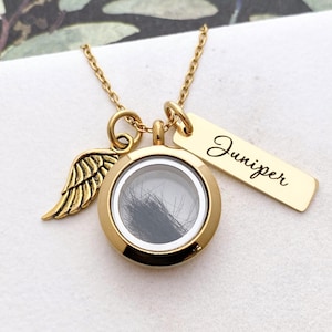 May include: A gold memorial necklace featuring a round locket with hair inside, a wing charm, and a tag engraved with "Juniper". The necklace hangs on a fine chain.