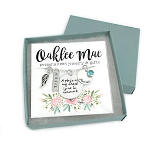 May include: A silver necklace with a charm that reads "A piece of my heart lives in heaven" and a charm with the date "8-7-16". The necklace also has a small angel wing charm and a charm with the name "Maxine". The necklace is in a small gift box with a white card that reads "Oaklee Mae personalized jewelry & gifts".