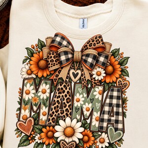 Peut inclure: Un sweat-shirt couleur crème avec une impression graphique du mot MAMA en grandes lettres. Les lettres sont décorées de motifs de marguerites, d'imprimé léopard et de carreaux vichy noirs et blancs. Un grand nœud, des tournesols et des cœurs entourent le texte.