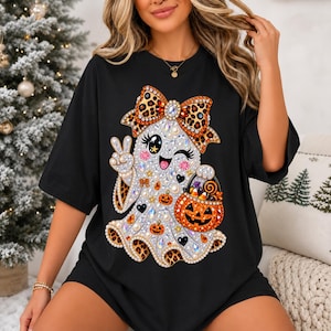 Peut inclure: Un t-shirt noir à manches courtes orné d'un motif central représentant un fantôme décoré de strass et de perles. Le fantôme porte un nœud à motif léopard, fait un signe de paix et tient un seau citrouille rempli de bonbons et de petites citrouilles.