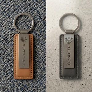 May include: Two rectangular keychains with silver metal rings. The left keychain is tan with a silver plate engraved with a compass icon and the coordinates 41.8781 N, 87.6298 W. The right keychain is grey with a silver plate engraved with a heart icon and the word CONNECTED.