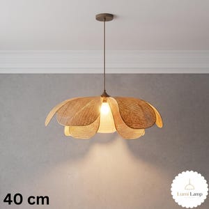 May include: A pendant light fixture with a woven tan rattan shade shaped like a flower with multiple petals. The lamp hangs from a thin brown cord and emits a warm light from a central bulb. The text 40 cm and Lumi Lamp are visible.