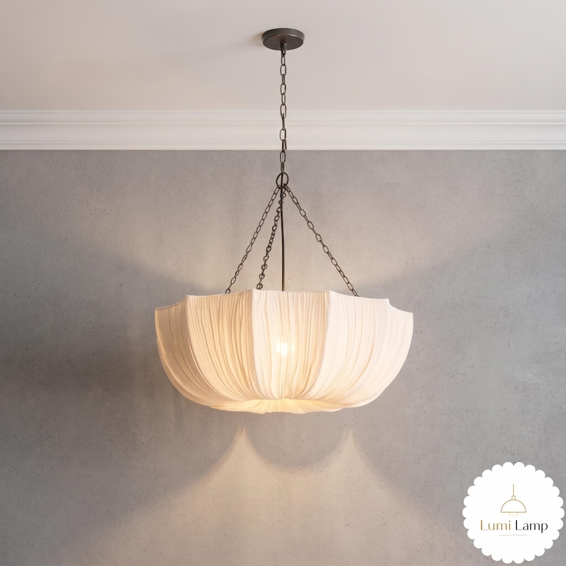 May include: A pendant light fixture featuring a wide, off-white fabric shade with vertical pleats. The shade is suspended from a dark metal ceiling canopy by three thin chains that meet at a central rod, creating a bowl-like, inverted umbrella shape.