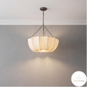 May include: A pendant light fixture featuring a cream-colored fabric shade with a scalloped, umbrella-like design. The shade is suspended by a dark metal chain and canopy. The light emits a warm glow from the center, with the text Lumi Lamp.