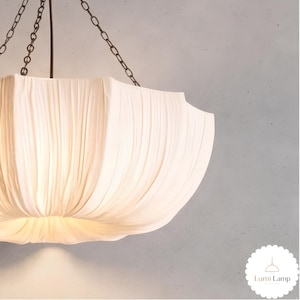 May include: A white pendant light fixture with a pleated, fabric-like shade suspended by dark metal chains. The shade has a wide, bowl-like shape with vertical folds. A small logo in the bottom right corner reads Lumi Lamp with a line drawing.