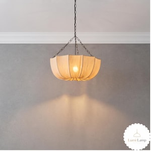 May include: A beige fabric pendant light with a scalloped, umbrella-like shade suspended by a dark metal chain. The light emits a warm glow from a central bulb, casting a soft downward light. A small white circular logo reads Lumi Lamp.