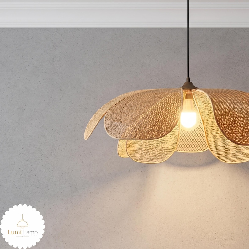 May include: A pendant light fixture featuring a woven tan rattan shade shaped like a flower with multiple overlapping petals. The lamp hangs from a thin black cord and contains a single glowing light bulb at its center. The logo Lumi Lamp is visible.