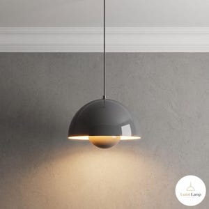 May include: A glossy dark gray dome-shaped pendant lamp suspended by a thin cord. The fixture features a smaller, centered inner dome that emits a warm, soft glow from beneath the main shade. The text Lumi Lamp is visible in the corner.