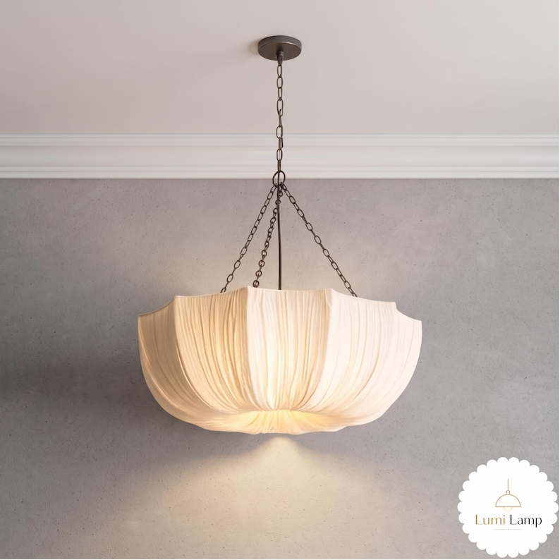 May include: A pendant light fixture featuring a large, off-white fabric shade with vertical pleats. The shade is suspended by a dark metal chain assembly connected to a matching circular ceiling canopy. The Lumi Lamp logo is visible in the corner.